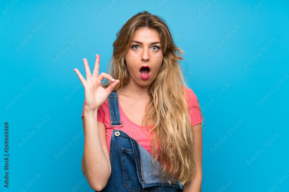 Young blonde woman with overalls over isolated blue wall surprised and showing ok sign