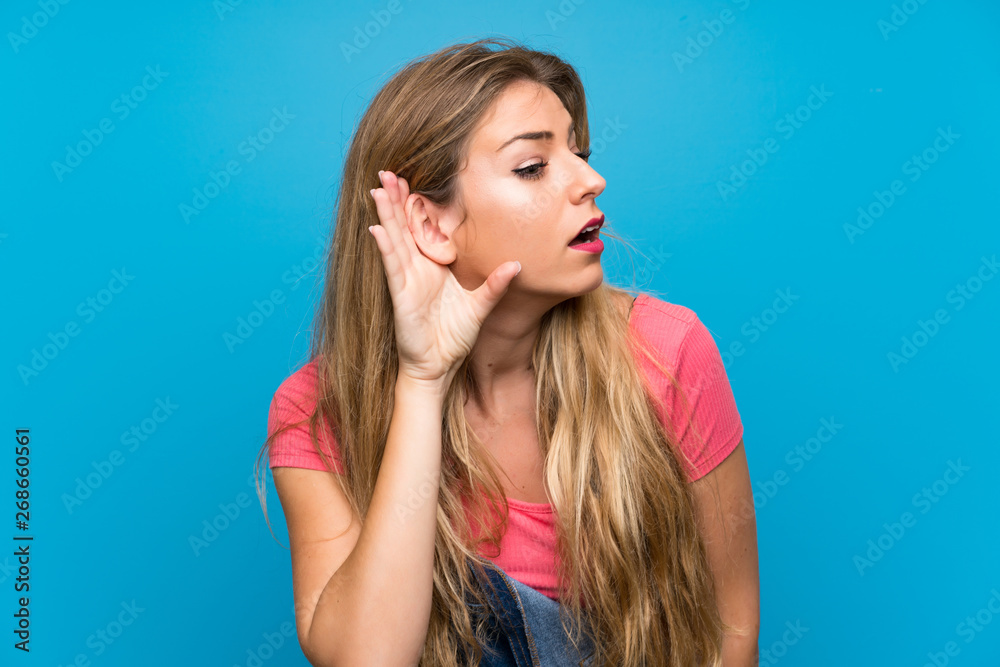 Young blonde woman with overalls over isolated blue wall listening to something by putting hand on the ear Young blonde woman with overalls over isolated blue wall listening to something by putting hand on the ear