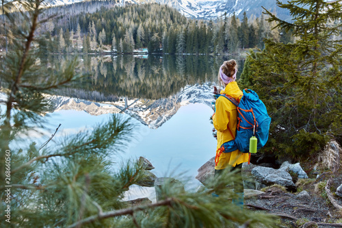 Outdoor shot of young traveler with bag, stands back to camera, enjoys mountains, fresh air and small lake, looks calmly at great spectacle of nature, stands near stones and firs, has vacation