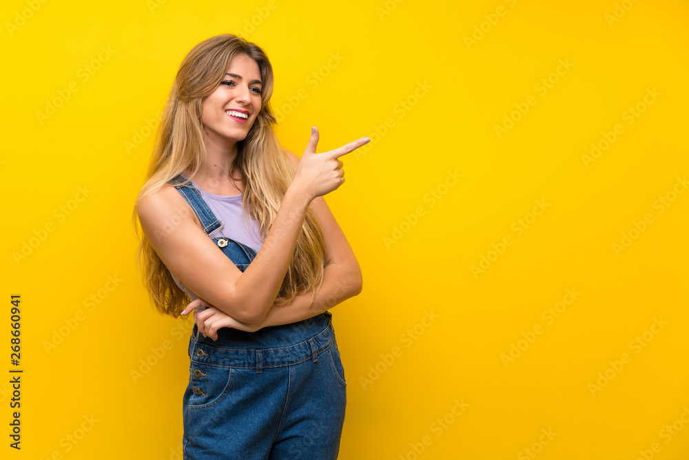 Young blonde woman with overalls over isolated yellow background pointing finger to the side Young blonde woman with overalls over isolated yellow background pointing finger to the side