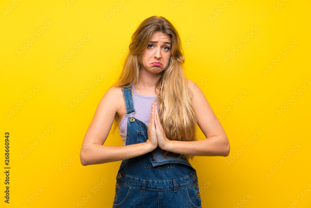 Young blonde woman with overalls over isolated yellow background pleading Young blonde woman with overalls over isolated yellow background pleading