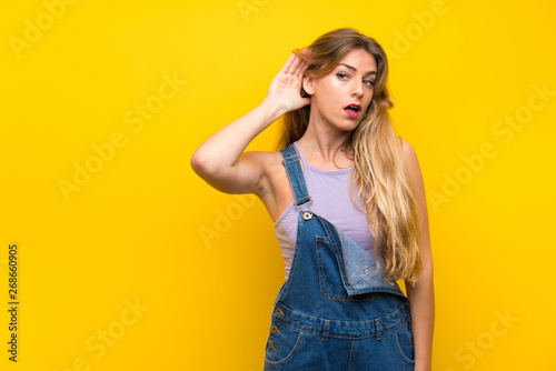 Young blonde woman with overalls over isolated yellow background listening something