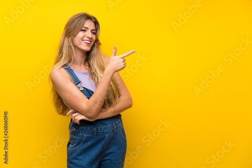 Young blonde woman with overalls over isolated yellow background pointing finger to the side