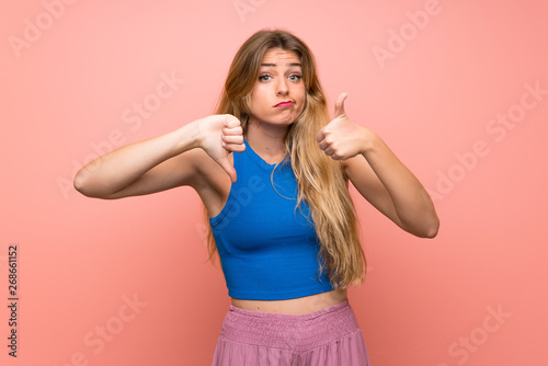 Young blonde woman over isolated pink background making good-bad sign. Undecided between yes or not