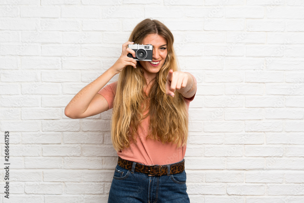 Young blonde woman over white brick wall holding a camera Young blonde woman over white brick wall holding a camera