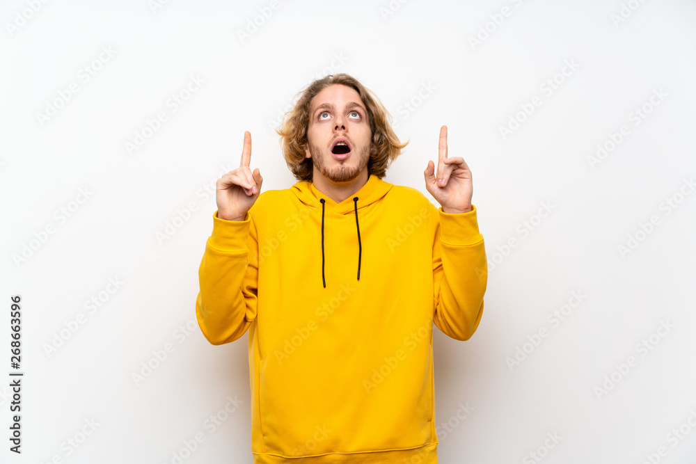 Blonde man with sweatshirt over white wall surprised and pointing up Blonde man with sweatshirt over white wall surprised and pointing up