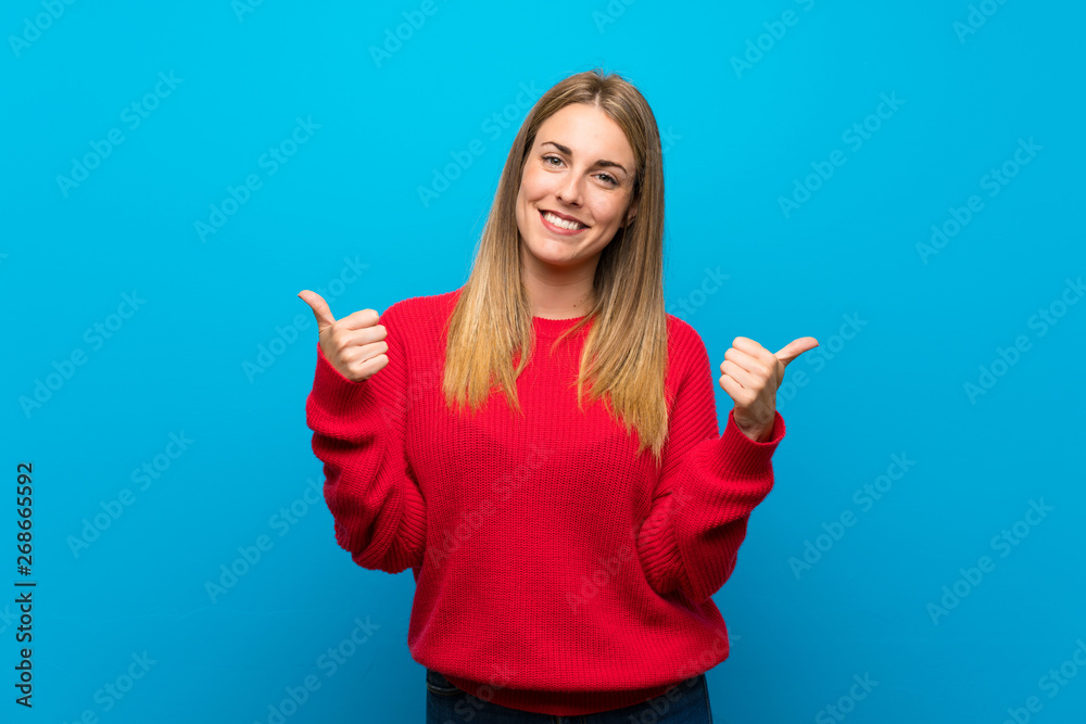 Woman with red sweater over blue wall with thumbs up gesture and smiling Woman with red sweater over blue wall with thumbs up gesture and smiling