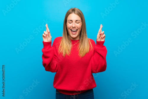 Woman with red sweater over blue wall with fingers crossing