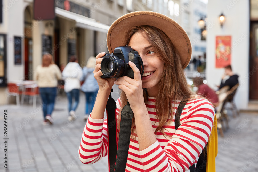 Professional female photographer uses photocamera for making pictures, takes photo of beautiful sights, dressed in stylish apparel against urban setting, spends free time in city, has journey