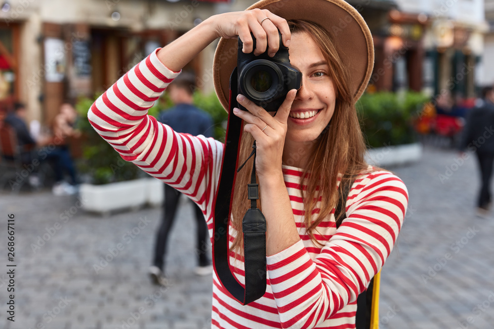 Happy female spends free time on hobby, takes picture of city street on camera during leisure, enjoys weekend travelling, wears headgear and striped jumper, strolls outdoors. People and lifestyle