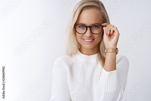 Charming female wants help pick glasses. Portrait of elegant and sensual young blond woman with daring and cheeky smile looking confident touching frames as posing against white background