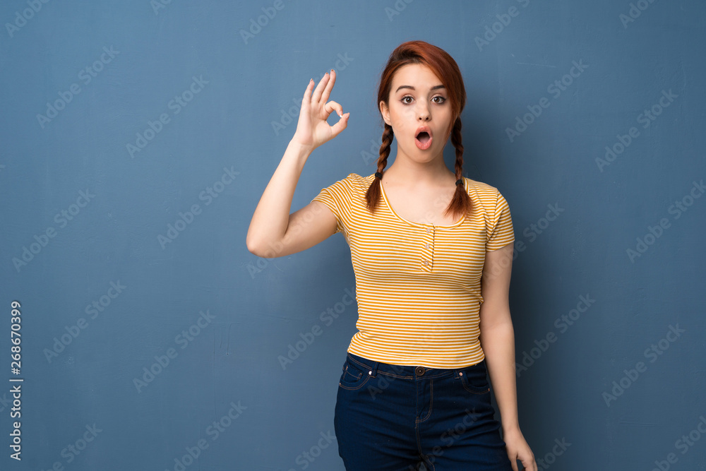 Young redhead woman over blue background surprised and showing ok sign Young redhead woman over blue background surprised and showing ok sign