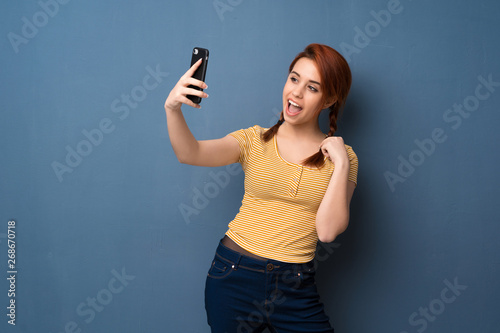 Young redhead woman over blue background making a selfie