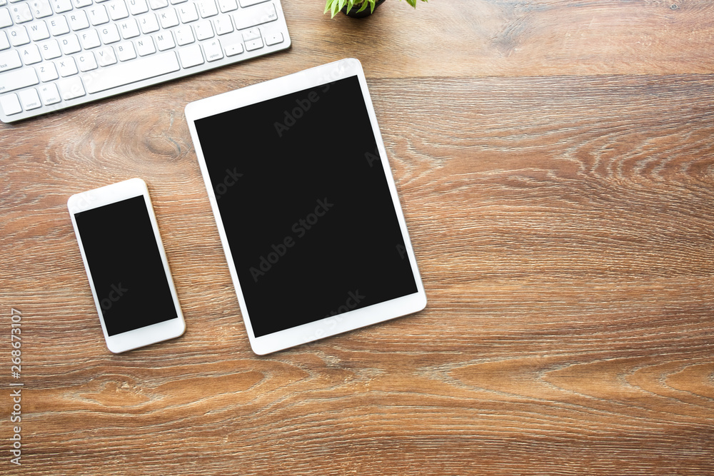 White smartphone and table with blank mockup screens are on top of wood office desk table with computer keyboard. Top view with copy space, flat lay. White smartphone and table with blank mockup screens are on top of wood office desk table with computer keyboard. Top view with copy space, flat lay.