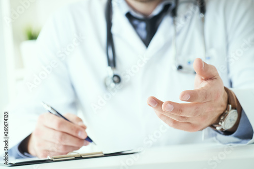 Male doctor making welcome gesture, politely inviting patient to sit down in medical office. Photo with depth of field.