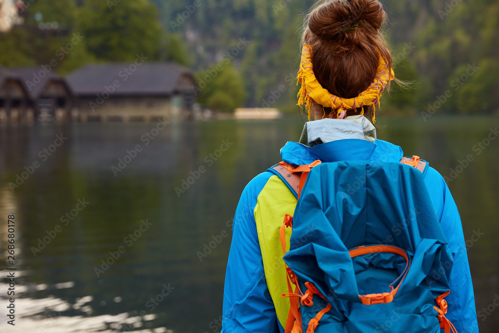 People, recreation time concept. Female tourist stands back to camera carries backpack, dressed in casual jacket focused into distance with small house in background admires beatiful river nature view