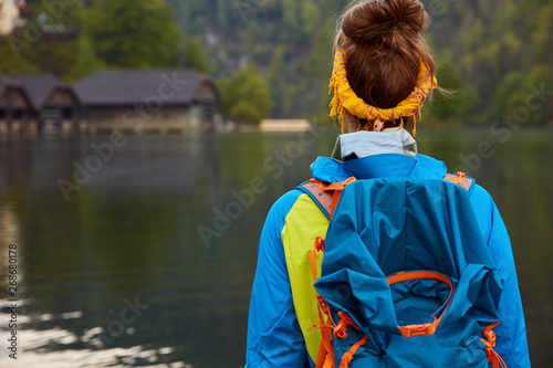 People, recreation time concept. Female tourist stands back to camera carries backpack, dressed in casual jacket focused into distance with small house in background admires beatiful river nature view