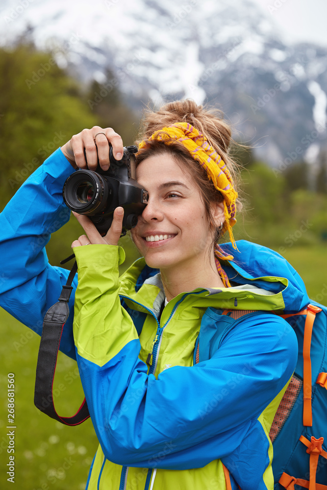 Φωτογραφία Female nature photographer uses camera for making photos of awesome mountain view, shoots during stroll, wears yellow scarf on head and anorak, green plantation in background
