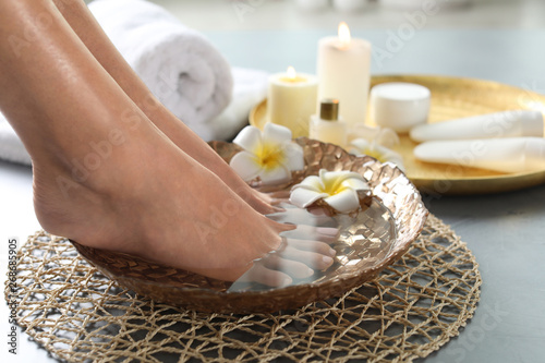 Closeup view of woman soaking her feet in dish with water and flowers on grey floor. Spa treatment