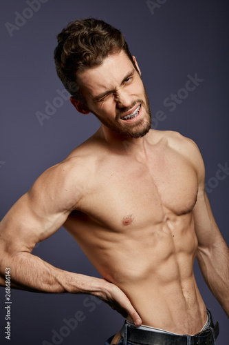 unhappy shirtless muscular man in jeans is holding his pain back. sportsman has problems with back, man with hand on his hip expresses painful emotion