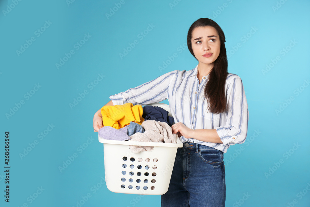 Displeased young woman holding basket with laundry on color background Displeased young woman holding basket with laundry on color background