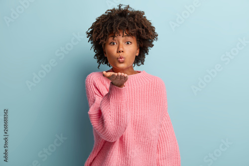 Glamorous dark skinned woman sends air kiss, keeps lips folded, has Afro haircut, wears casual pink jumper, isolated over blue background, says be valentine. Flirting and body language concept