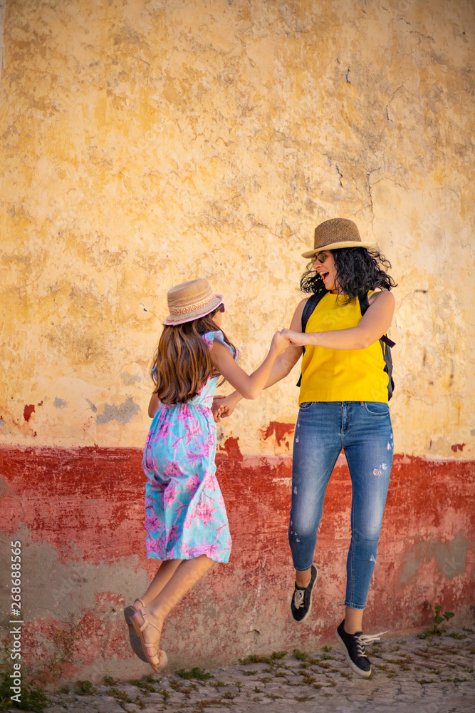 A mother and her daughter is jumping in front of a cracked ( rustic ) wall with happiness