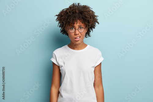 Half length shot of dissatisfied female smirks face, reacts on something unpleasant, wears casual clothes, spectacles, isolated on blue background, expresses dissatisfaction, doesnt like bad treament