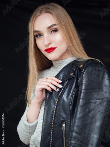 Fashion portrait stylish pretty blonde woman with bright trandy makeup red lips posing outdoor black backgrpound wearing a rock black style leather jacket on her shoulder having fun in city.