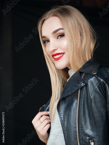Fashion portrait stylish pretty blonde woman with bright trandy makeup red lips posing outdoor black backgrpound wearing a rock black style leather jacket on her shoulder having fun in city.
