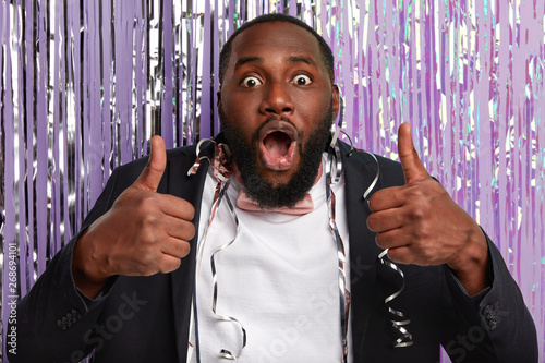 Shocked ethnic man opens mouth from wonder, stares at camera, raises thumbs up, fascinated by something awesome, demonstrates okay gesture, wears white shirt and black suit, serpentine everywhere photo