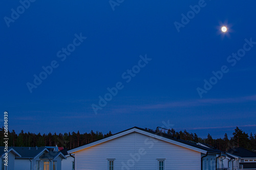 Gorgeous view of the natural landscape on a summer night. Roofs of villas, forest, moon, blue sky and clouds converging on the horizon. Beautiful colorful nature background. © Alex