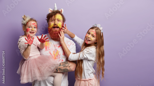 Emotive single father spends free time with daughters, paint faces, use watercolors, two kids have joyful expression, play with dad. Redhead family have fun, isolated over purple wall, copy space