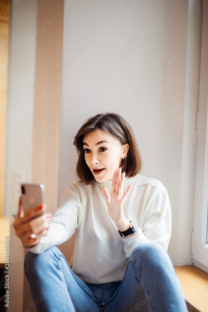 Happy woman sitting on the windowsill and making selfie on smartphone while waving at camera Happy woman sitting on the windowsill and making selfie on smartphone while waving at camera