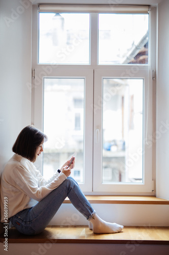 Happy woman reading positive news on web page while browsing internet on smartphone. Successful female blogger sitting on windowsill and tying sms message. Shopping online enjoying discounts