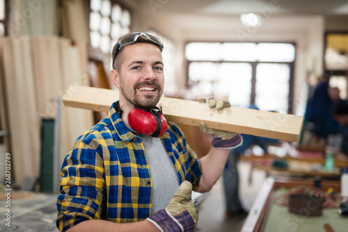 Happy smiling carpenter holding wood material and thumbs up in his carpentry workshop.