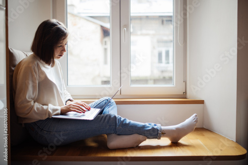 Fotografi Beautiful young woman sitting on windowsill with laptop computer.