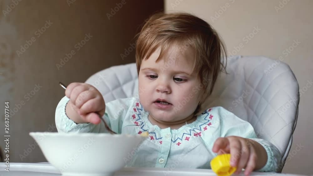 Little cute baby girl sits in a soft high chair and independently eats a spoon of pasta close-up in slow motion. Elbow macaroni falling from a child spoon. Kid holds in his hand a bright toy. Little cute baby girl sits in a soft high chair and independently eats a spoon of pasta close-up in slow motion. Elbow macaroni falling from a child spoon. Kid holds in his hand a bright toy.