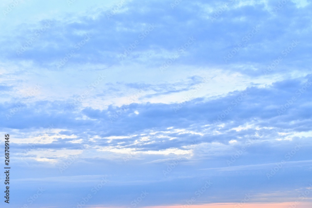 Gentle blue sky with a pink thin strip of sunset in the lower right corner. Bright blue clouds stretched horizontally and slightly diagonally upwards to the right, leaving bright celestial openings.
