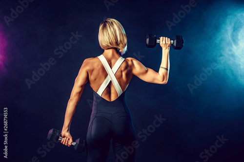 Back view of a sporty woman in fashionable sportswear does the exercises with dumbbells. Photo of muscular woman on dark background with smoke. Strength and motivation.