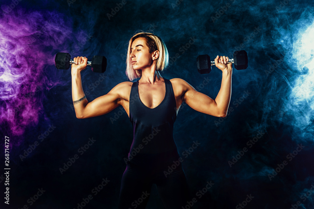 Attractive sporty blonde woman in fashionable sportswear does the exercises with dumbbells. Photo of muscular woman on dark background with smoke. Strength and motivation. Attractive sporty blonde woman in fashionable sportswear does the exercises with dumbbells. Photo of muscular woman on dark background with smoke. Strength and motivation.