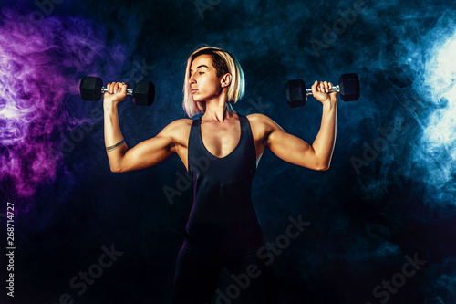 Attractive sporty blonde woman in fashionable sportswear does the exercises with dumbbells. Photo of muscular woman on dark background with smoke. Strength and motivation.