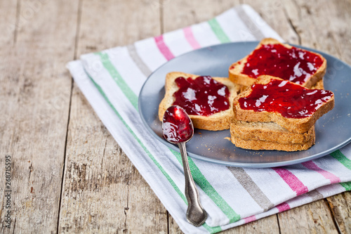Fresh toasted cereal bread slices with homemade cherry jam and spoon on ceramic plate closeup on linen napkin on rustic wooden table background.