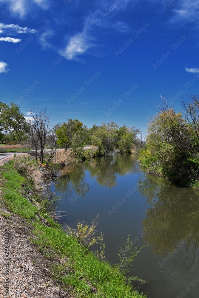 Jordan River Parkway Trail, Redwood Trailhead bordering the Legacy Parkway Trail, panorama views with surrounding trees and silt filled muddy water along the Rocky Mountains, Salt Lake City, Utah. 