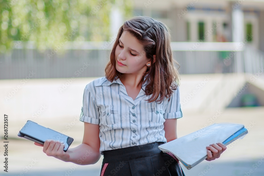 Obraz premium Girl schoolgirl compares the tablet and textbooks. The child is holding a tablet and books. The student has an idea. The girl in the shirt opposite the school.
