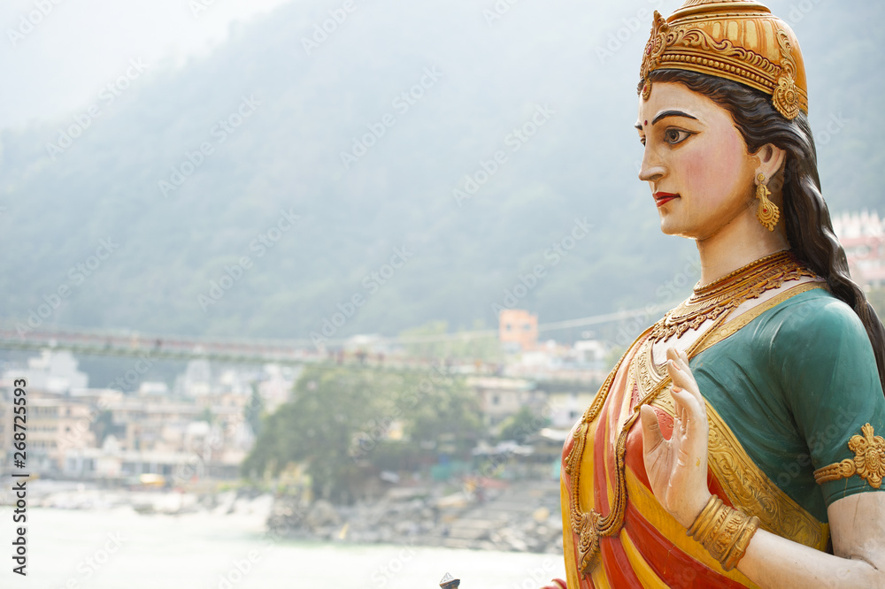 Close-up view of the statue of sitting Goddess Parvati on the riverbank of the Ganges river. Blurred Lakshman Jhula bridge in the background. Rishikesh, Uttarakhand, India.
