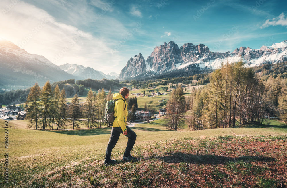 Obraz premium Young man in yellow jacket with backpack walking on the green meadow and looking on mountains at sunset in spring. Landscape with sporty guy, snowy rocks, trees, houses, blue sky in Italy. Travel