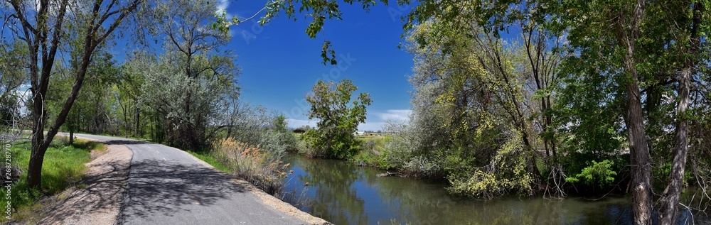 Fototapeta premium Jordan River Parkway Trail, Redwood Trailhead bordering the Legacy Parkway Trail, panorama views with surrounding trees and silt filled muddy water along the Rocky Mountains, Salt Lake City, Utah. 