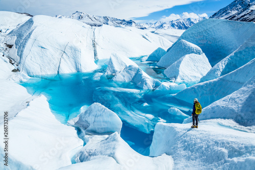 Young woman standing near deep blue lake on the Matanuska Glacier in Alaska. She wears a backpack and helmet with ice axe in hand for summer glacier travel. photo