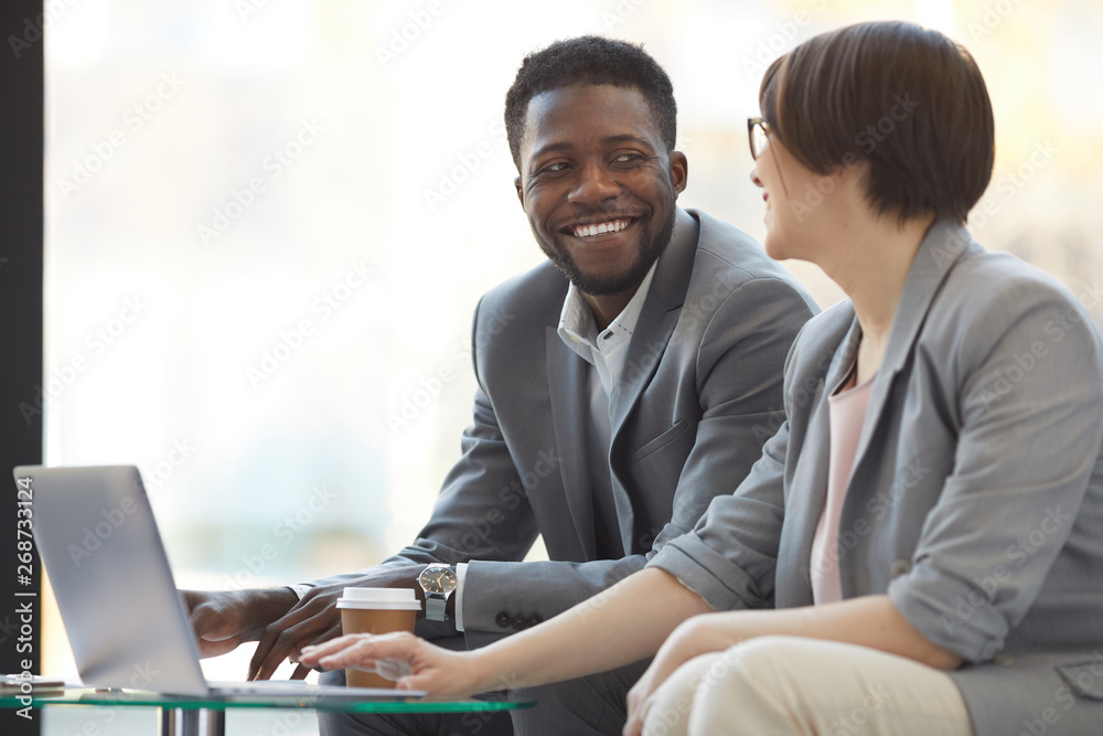 Positive creative multi-ethnic business team sitting in lobby and looking at each other while discussing sales information on laptop